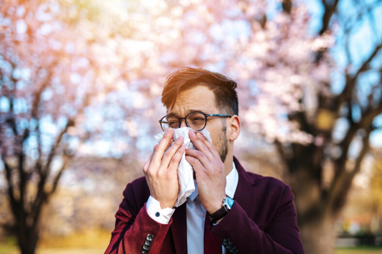 Heuschnupfen.jpg Ein Mann in einem Anzug benutzt ein Taschentuch, um sich die Nase zu putzen, vor blühenden Bäumen im Hintergrund.