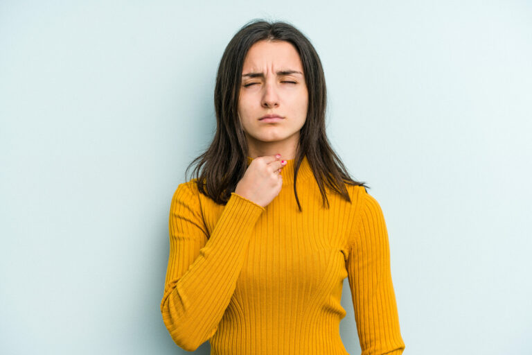 Frau in gelbem Pullover, die nachdenklich auf ihre Kehle zeigt, vor einer hellblauen Wand.