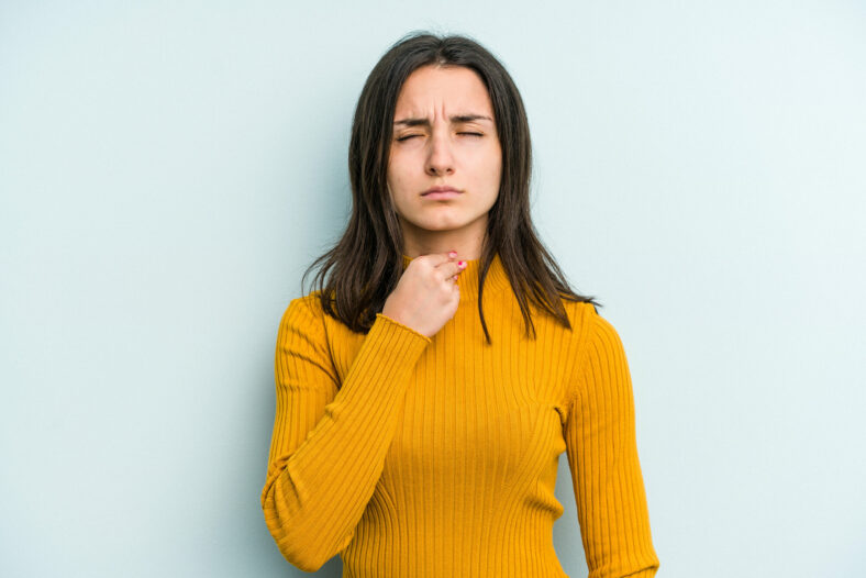 Frau in gelbem Pullover, die nachdenklich auf ihre Kehle zeigt, vor einer hellblauen Wand.