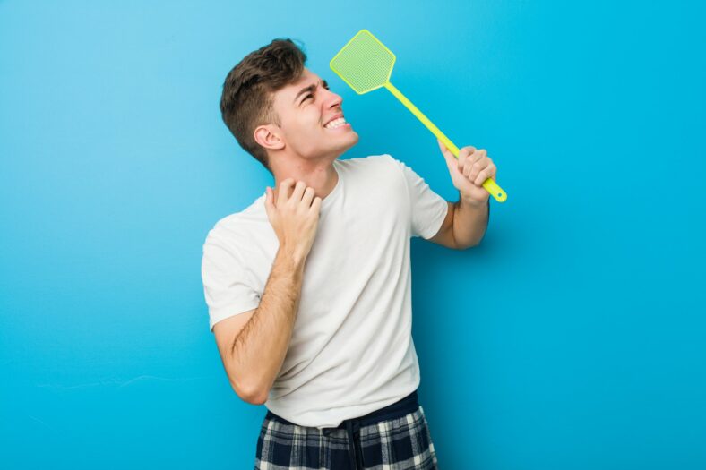 plagegeister.jpg Junger Mann in einem weißen T-Shirt hält ein grünes Insektenspray mit einem lächelnden Gesicht vor einer blauen Wand.