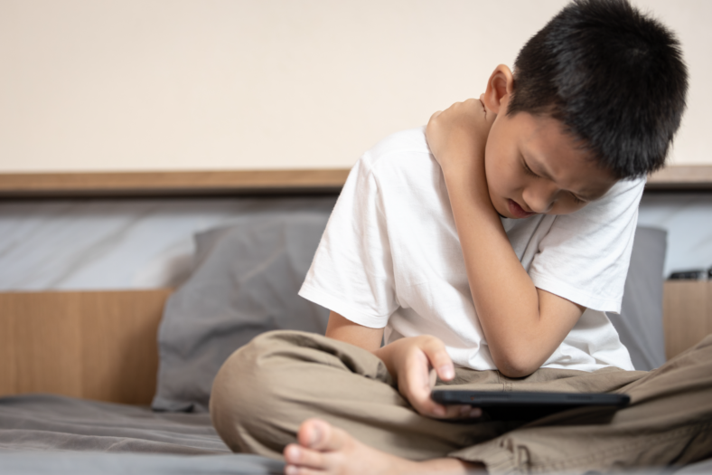 rueckenfit.png Ein Junge sitzt auf einem Bett, hält ein Tablet und fasst sich schmerzverzerrt an den Hals.