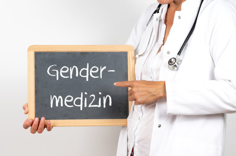 Eine Person in Arztkleidung hält eine Tafel mit der Aufschrift "Gendermedizin".