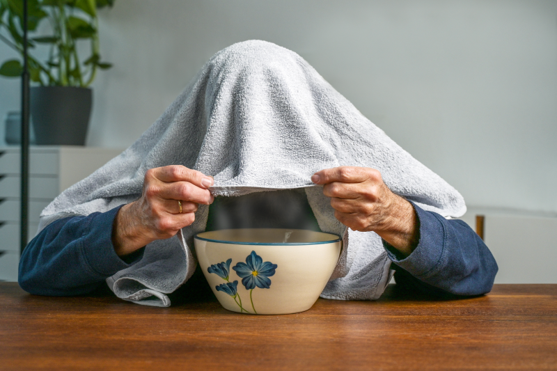 Dampfinhalation.png Eine Person mit einem Handtuch über dem Kopf hält eine Schüssel mit Blumenmuster auf einem Tisch.