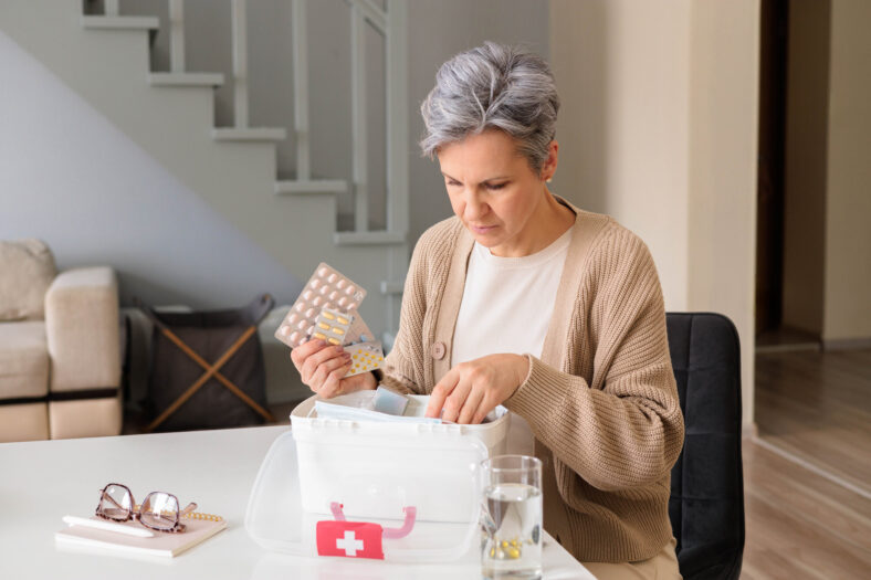 Eine Frau in einem hellen Raum untersucht einen durchsichtigen Medikamentenbehälter und hält eine Packung Tabletten.