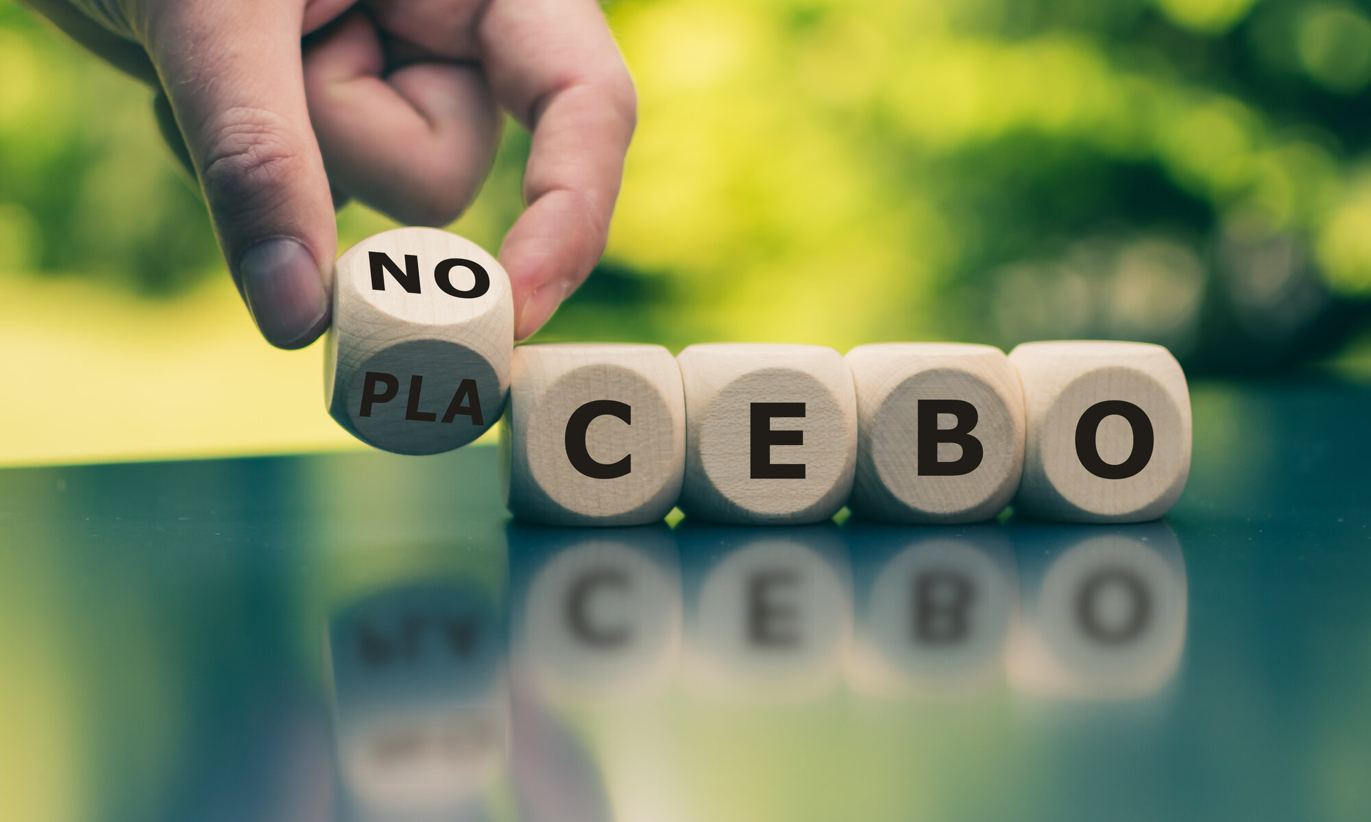 placebo-nocebo.jpeg Holzwürfel mit Buchstaben, die das Wort 'NO PLACEBO' bilden, auf einem Tisch in der Natur.